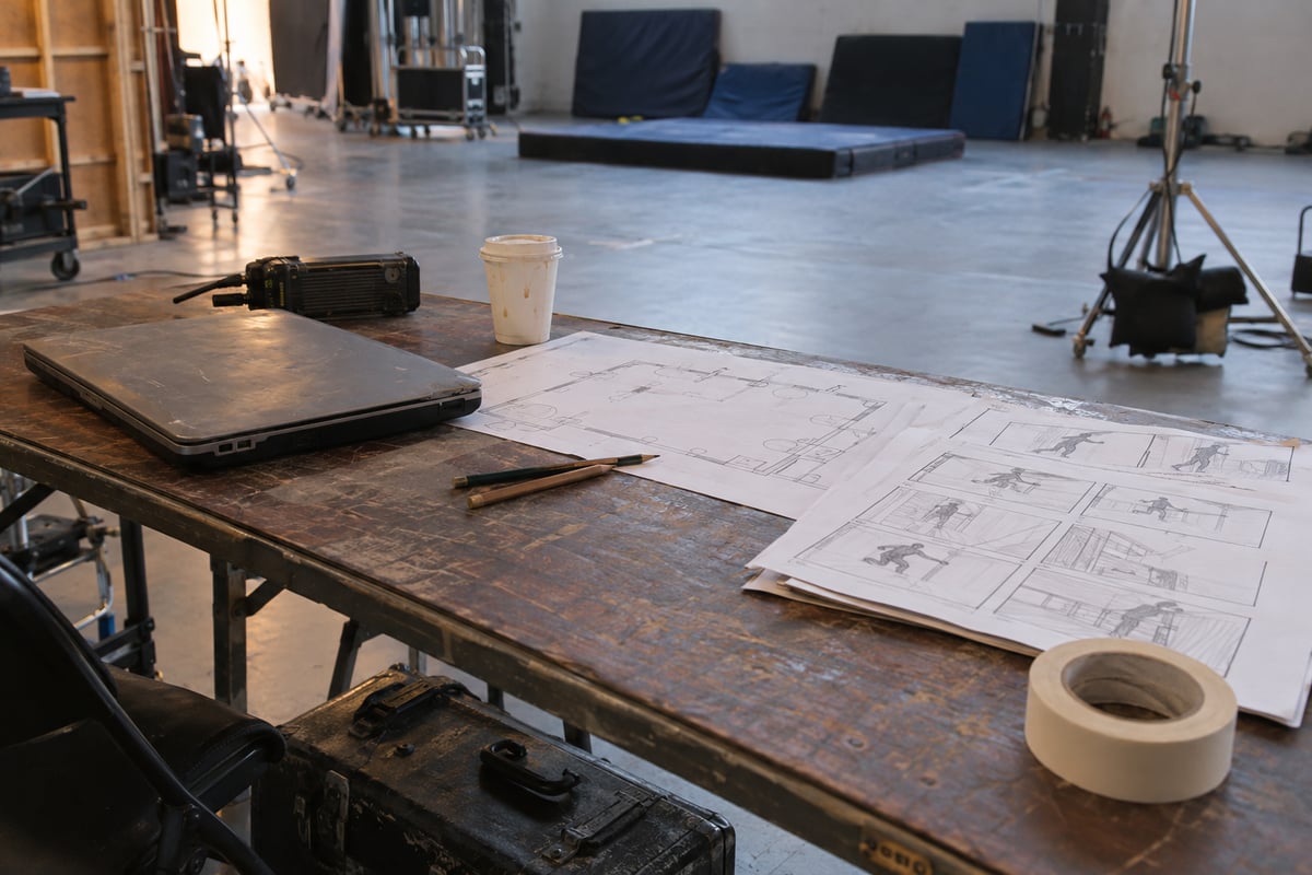 Storyboards, a floor plan, radio and worn laptop on a work table beside a rehearsal floor.